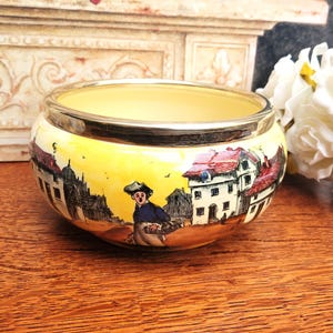 May include: A decorative ceramic bowl with a yellow interior and a silver rim. The exterior features a painted landscape scene with buildings, a figure, and a yellow and orange sky. The bowl is likely for decorative purposes.