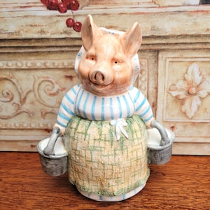 Puede incluir: Figura de cerámica de un cerdo con camisa a rayas azules y blancas, delantal con estampado verde y blanco y gorro blanco. El cerdo sostiene dos cubos grises. La figura tiene un aspecto detallado pintado a mano.