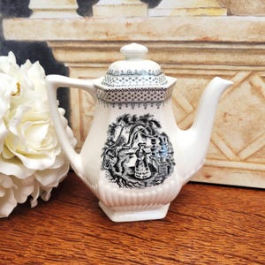 Peut inclure: Une théière en céramique blanche avec une scène pastorale en noir et blanc. La théière présente un bec courbé, une anse et un couvercle avec un motif décoratif. Le motif comprend une femme près d'un arbre et d'un bâtiment. La théière est sur une surface en bois.
