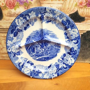 Op de afbeelding: Een blauw-witte verdeelde serveerschaal met een bloemen- en pastorale ontwerp. De schaal heeft een centrale scène van een paardenkoets en een bloemenrand. De schaal wordt ondersteund door een witte standaard.