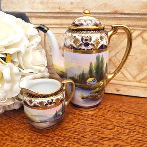 May include: A vintage porcelain coffee set featuring a tall coffee pot and a small creamer. The set is painted with a landscape scene and accented with gold trim. The coffee pot measures approximately 10 inches in height.