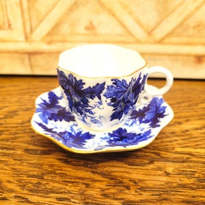 May include: A white teacup and saucer set with a blue floral pattern and gold trim. The teacup has a unique, scalloped rim and a small handle. The saucer mirrors the teacup's design, creating a cohesive and elegant look.
