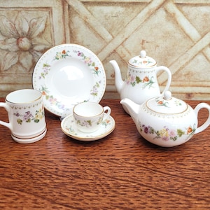 Może przedstawiać: Biały porcelanowy zestaw do herbaty z kwiatowymi wzorami. Zestaw zawiera czajniczek, filiżankę ze spodkiem, kubek i małą miseczkę. Krawędzie są wykończone złotem. Kwiatowy wzór obejmuje różowe, żółte i fioletowe kwiaty.