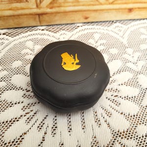 May include: A black round box with a gold embossed image of a person holding a knife. The box is on a white lace surface.