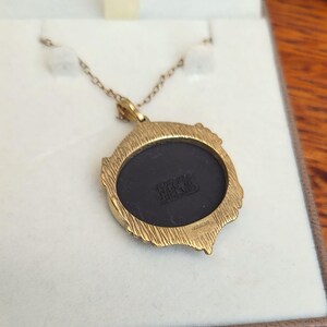 May include: A gold-toned pendant necklace with a round, black center. The pendant has a textured, ornate frame. The chain is delicate and gold-colored. The back of the pendant has the text "WRIGHT & ENGLAND".