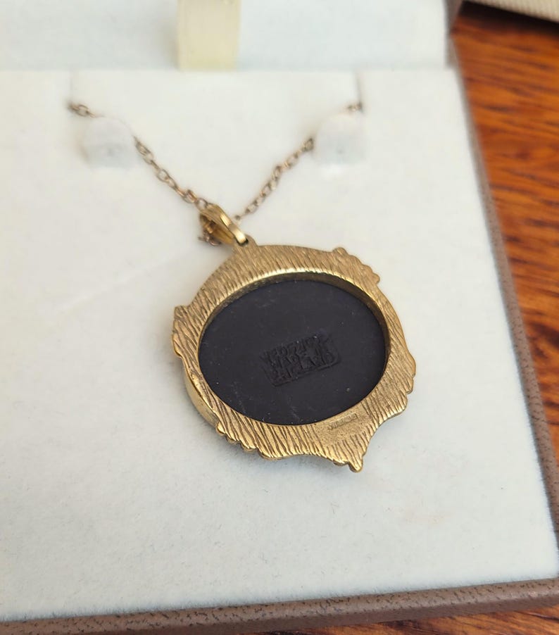 May include: A gold-toned pendant necklace with a round, black center. The pendant has a textured, ornate frame. The chain is delicate and gold-colored. The back of the pendant has the text "WRIGHT & ENGLAND".