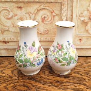 May include: A pair of white ceramic salt and pepper shakers with a floral design. The shakers have a gold rim and base, and are decorated with colorful flowers, including pansies, roses, and blue cornflowers. The shakers are approximately 4 inches tall.