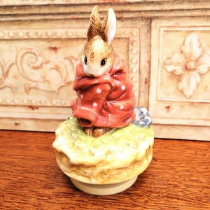 Peut inclure: Figurine en céramique représentant un lapin vêtu d'un manteau rouge à pois blancs. Le lapin est assis sur un socle jaune et vert. Objet de collection, il est décoratif et charmant.