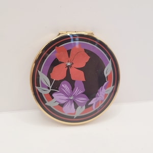 May include: A round, gold-toned compact mirror with a black background and a floral design. The flowers are red, purple, and gray.
