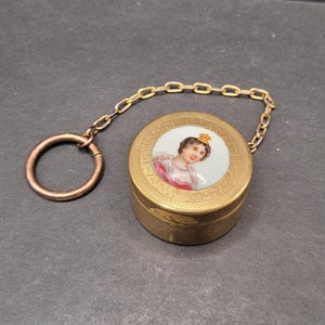 Peut inclure: Petite boîte ronde en métal doré avec un portrait de femme sur le couvercle. Le portrait est dans un cercle blanc et représente une femme couronnée. Une chaîne et un anneau dorés sont attachés à la boîte.