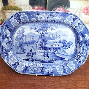 Peut inclure: Plat à servir en céramique bleu et blanc avec une scène de paysage. Le plat a une bordure détaillée de fleurs et de feuilles. L'image centrale représente un pont, des bâtiments et des personnages à cheval.