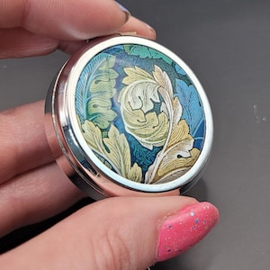 May include: A round silver locket featuring a colourful floral design. The design incorporates swirling leaves in shades of green, blue, and yellow.