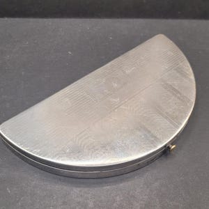 May include: A silver-colored, semi-circular box with a hinged lid. The lid and base have a textured, engraved pattern. The box is closed with a small, gold-colored clasp. The box is on a dark surface.