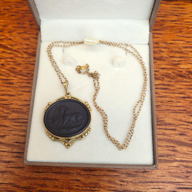May include: A gold-toned necklace with a black oval pendant featuring a carved image of a horse. The pendant is set in a decorative gold frame. The necklace is presented in a brown box with a white interior, set against a wooden surface.