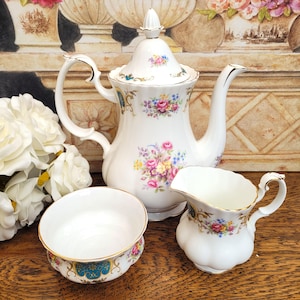 Puede incluir: Un juego de té de porcelana blanca con diseños florales. El juego incluye una tetera con tapa, una jarra de crema y un cuenco pequeño. Los bordes de cada pieza están adornados con detalles dorados. El estampado floral presenta flores rosas, amarillas y azules.