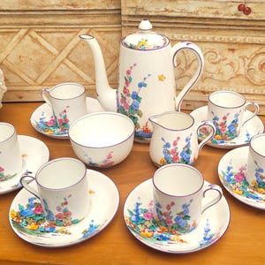 Peut inclure: Service à thé en porcelaine blanche avec des motifs floraux. L'ensemble comprend une théière, des tasses, des soucoupes, un pot à crème et un sucrier. Les fleurs sont bleues, roses et jaunes. L'ensemble est disposé sur une surface en bois.