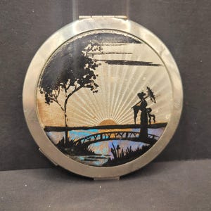 May include: A silver compact mirror featuring a black silhouette of a woman with an umbrella standing on a bridge over a lake. The background depicts a sunset and a tree. The mirror has an ornate design around the edge.