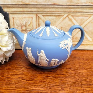 May include: A light blue ceramic teapot with white relief detailing. The teapot features a lid with a knob, a curved handle, and a spout. The relief depicts classical figures and decorative elements. The teapot is displayed on a wooden surface.