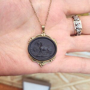 May include: A gold-toned necklace with a black oval pendant featuring a carved horse design. The pendant is framed by a decorative gold border. A gold ring with diamond-shaped accents is also visible.