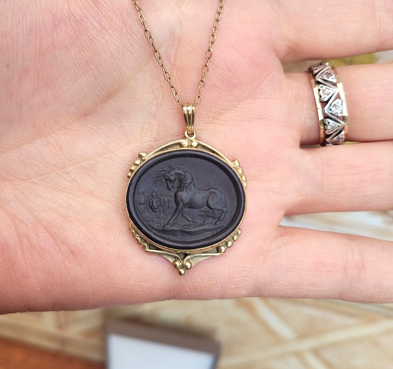 May include: A gold-toned necklace with a black oval pendant featuring a carved horse design. The pendant is framed by a decorative gold border. A gold ring with diamond-shaped accents is also visible.