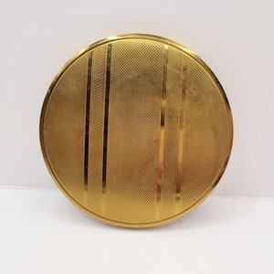 May include: A round, gold-colored compact with a textured, patterned design. The compact has a raised rim and a smooth, shiny surface.