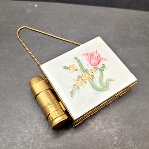Peut inclure: Un compact vintage en or avec un motif floral représentant une tulipe rose et des fleurs jaunes. Le compact est doté d'une chaîne et d'un tube de rouge à lèvres.