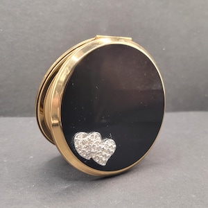 Op de afbeelding: Een vintage compacte spiegel met een zwarte emaille afwerking en een goudkleurig frame. Twee met strass bezette harten sieren de voorkant. De ronde compact is gesloten en toont zijn elegante ontwerp.