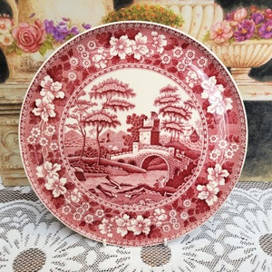 May include: A vintage red and white ceramic plate with a floral border and a central design of a bridge over a river with trees and a building in the background.