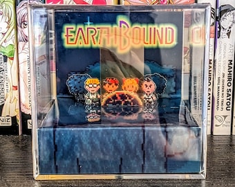 May include: A clear acrylic display case with a black and white pixelated image of the video game EarthBound. The case is sitting on a wooden shelf with other books and manga.