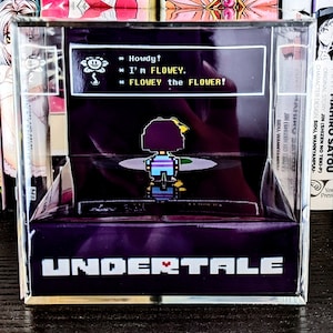 May include: A purple and black box with a clear front panel. The box features a pixelated image of a character from the video game Undertale. The text "UNDERTALE" is printed on the front of the box. The text inside the box reads "Howdy! I'm FLOWEY. FLOWEY the FLOWER!"