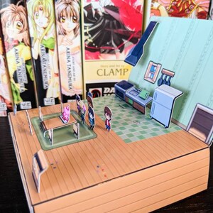 May include: A 3D papercraft model of a house with a kitchen interior. The model is made of cardboard and features a green tiled floor, a white fridge, and a table with chairs. There are also small figures of people standing around the table.