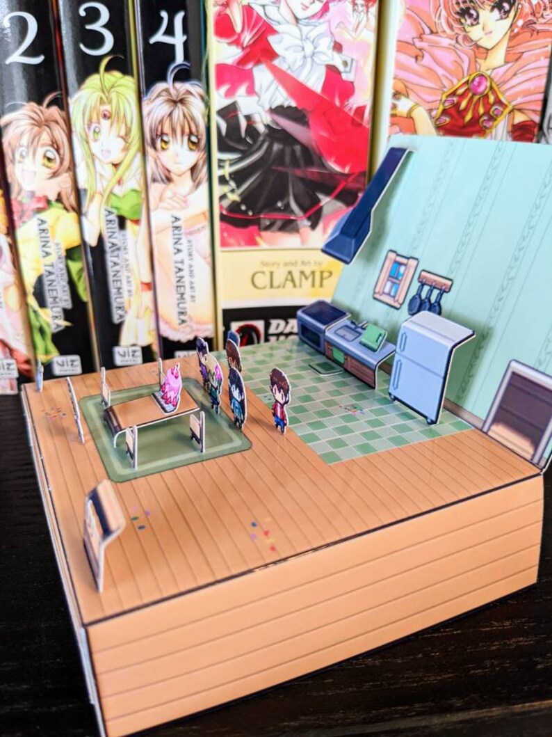 May include: A 3D papercraft model of a house with a kitchen interior. The model is made of cardboard and features a green tiled floor, a white fridge, and a table with chairs. There are also small figures of people standing around the table.