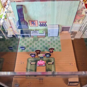 May include: A miniature diorama of a kitchen with a table set for a birthday party. Five pixelated figures stand around a table with a pink and white cake. The kitchen features a black stove, a sink, and a white refrigerator. The diorama is displayed in a clear acrylic box.