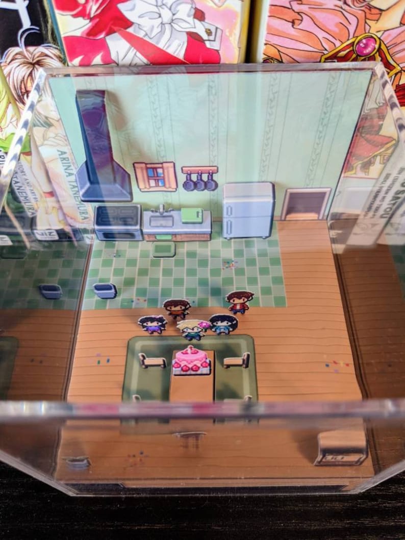 May include: A miniature diorama of a kitchen with a table set for a birthday party. Five pixelated figures stand around a table with a pink and white cake. The kitchen features a black stove, a sink, and a white refrigerator. The diorama is displayed in a clear acrylic box.