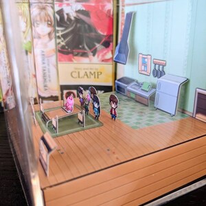 May include: A miniature papercraft diorama of a kitchen with a table and chairs, a stove, a refrigerator, and a sink. There are five small figures standing around the table. The diorama is displayed in a clear acrylic box.