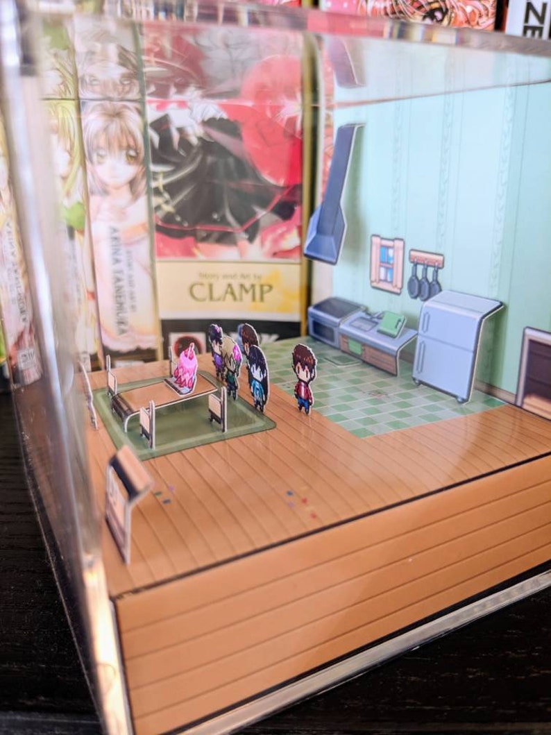 May include: A miniature papercraft diorama of a kitchen with a table and chairs, a stove, a refrigerator, and a sink. There are five small figures standing around the table. The diorama is displayed in a clear acrylic box.
