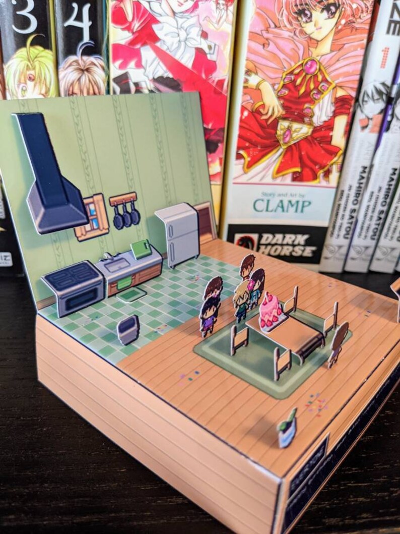 May include: A papercraft pop-up scene of a kitchen with a stove, sink, and fridge. Four figures are gathered around a table with a pink cake. The scene is set against a backdrop of manga books with the text "Story and Art by CLAMP DARK HORSE" visible.