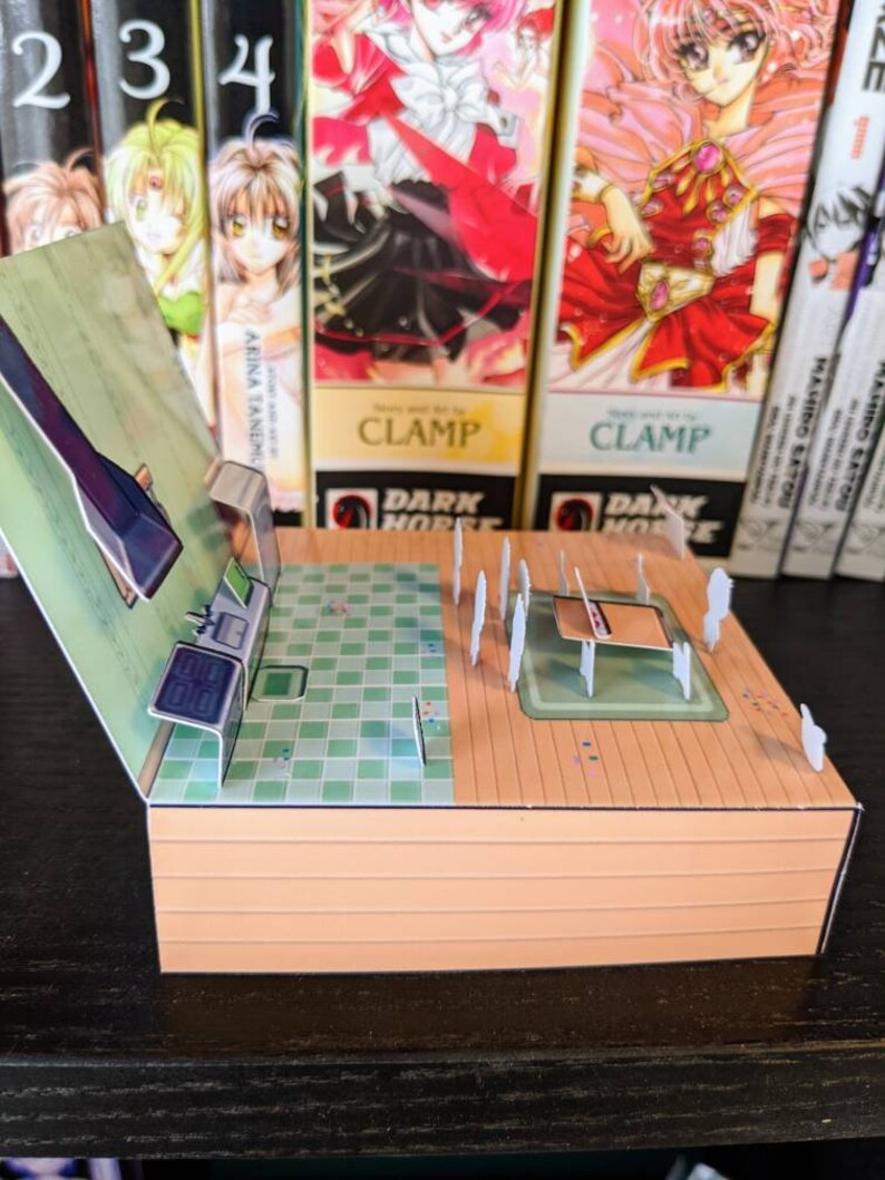 May include: A papercraft model of a room with a green tiled floor, a wooden floor, and a white countertop. The model is open and shows the interior of the room. The model is made of paper and cardboard and is painted in various colours.
