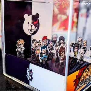 May include: A clear acrylic cube with a black and white background featuring a cartoon bear with a red eye. The cube contains a collection of small pixelated figures of characters from the Danganronpa video game series. The cube is sitting on a wooden surface.