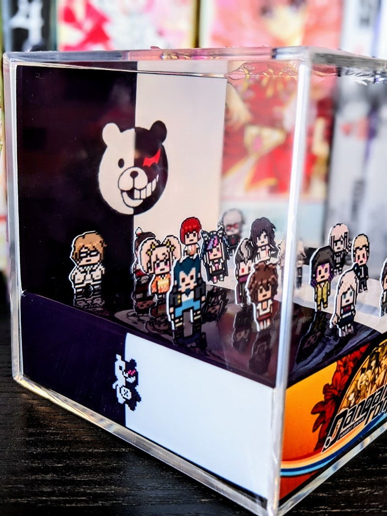 May include: A clear acrylic cube with a black and white background featuring a cartoon bear with a red eye. The cube contains a collection of small pixelated figures of characters from the Danganronpa video game series. The cube is sitting on a wooden surface.