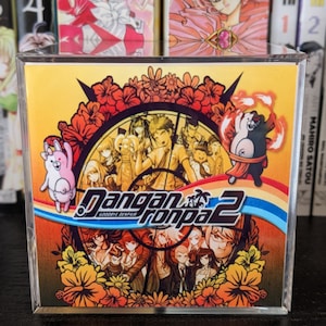 May include: A clear plastic case with a yellow and orange video game case inside. The case features a colorful illustration of characters from the video game "Danganronpa 2: Goodbye Despair".