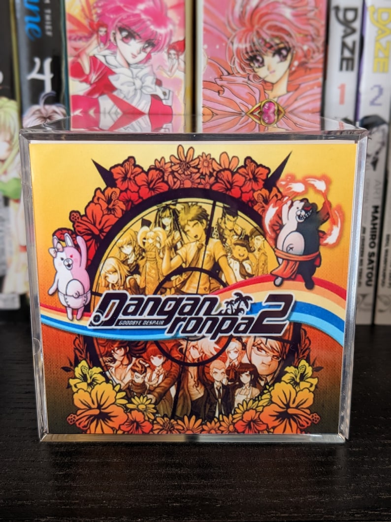 May include: A clear plastic case with a yellow and orange video game case inside. The case features a colorful illustration of characters from the video game "Danganronpa 2: Goodbye Despair".