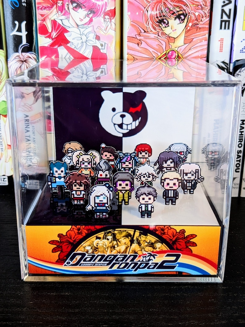 May include: A clear acrylic display case with a black and white bear mascot on the top. The case contains a collection of pixelated characters from the video game "Danganronpa 2: Goodbye Despair". The bottom of the case features a colorful image of the game's logo.