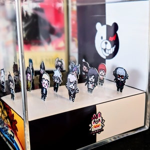 May include: A clear acrylic display case with a black and white background featuring a bear with a red eye. The case contains a collection of small pixelated figures of characters from the Danganronpa video game series.