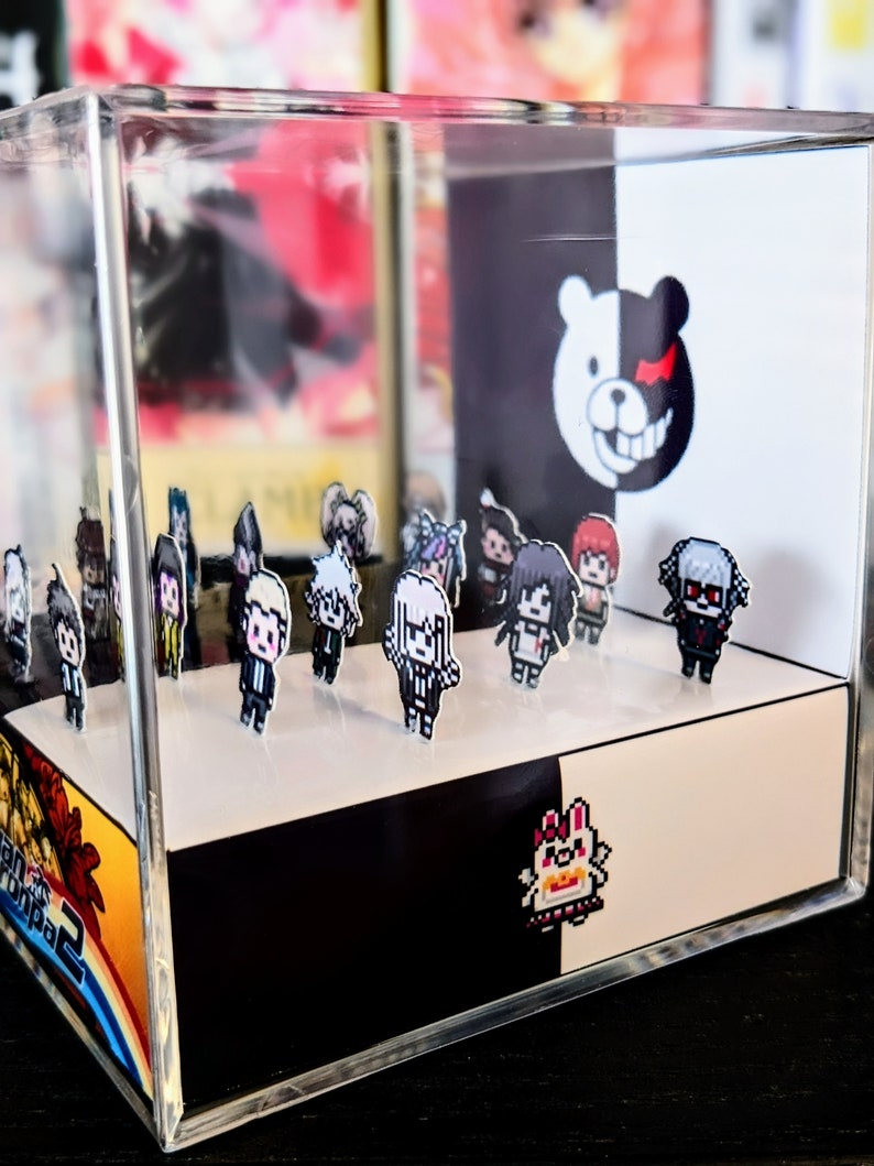May include: A clear acrylic display case with a black and white background featuring a bear with a red eye. The case contains a collection of small pixelated figures of characters from the Danganronpa video game series.