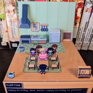 May include: A papercraft diorama depicting a birthday party with a cake, gifts, and characters. The diorama is made from cardboard and features a green and white background. The text "EVERYONE Happy birthday, dear BASIL! Happy birthday to youuuuu!" is printed on the bottom of the diorama.
