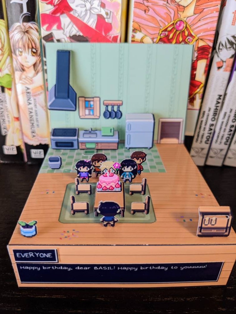 May include: A papercraft diorama depicting a birthday party with a cake, gifts, and characters. The diorama is made from cardboard and features a green and white background. The text "EVERYONE Happy birthday, dear BASIL! Happy birthday to youuuuu!" is printed on the bottom of the diorama.