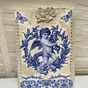 Puede incluir: Una placa de pared decorativa con un querubín sosteniendo un ramo de flores rodeado de una corona floral azul y blanca. La placa tiene un fondo blanco con un acabado desgastado y está decorada con mariposas y detalles florales.
