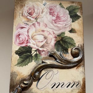 Könnte beinhalten: Rechteckige Holzplakette mit einem Blumendesign aus rosa Rosen und grünen Blättern. Eine dekorative, verzierte Metallrolle ist in der unteren rechten Ecke angebracht. Das Wort "Omm" ist in schwarzer Schrift geschrieben.