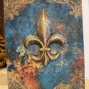 Puede incluir: Placa rectangular decorativa con un diseño de flor de lis. La placa presenta un fondo azul desgastado con detalles dorados y anaranjados. La flor de lis es en relieve, de color oro metálico, añadiendo un efecto tridimensional a la obra.